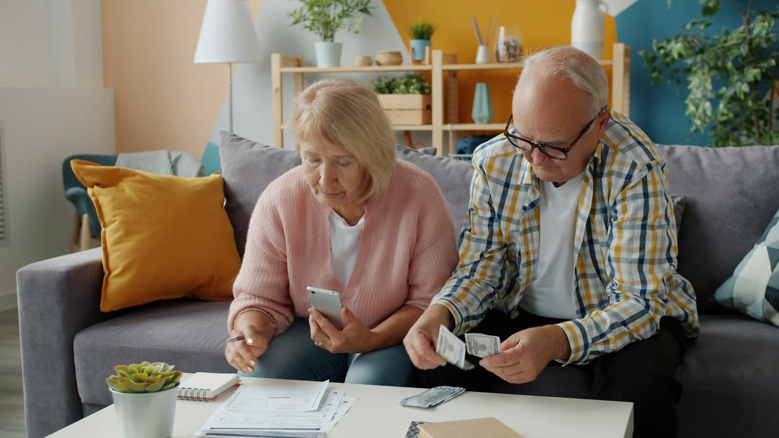 Older couple on a sofa counting cash and checking paperwork with a phone