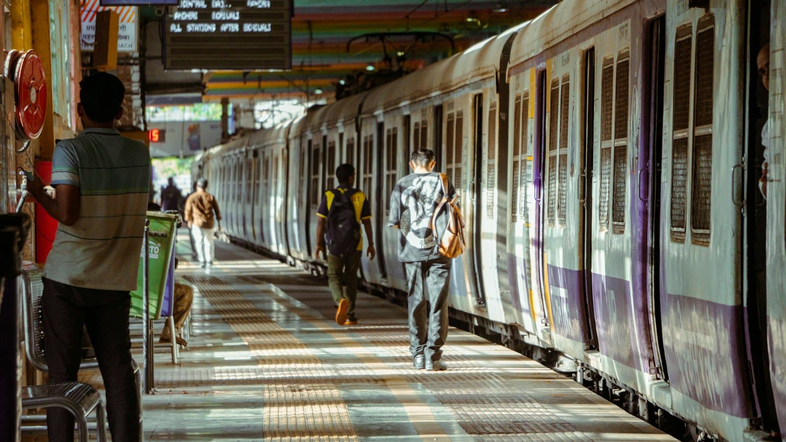 Young men with backpacks walking along a sunlit suburban train platform in Mumbai